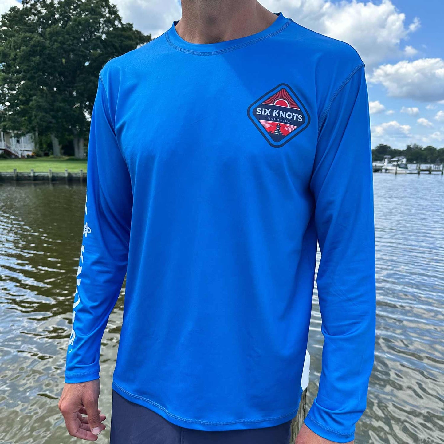 Man standing near the water wearing the Six Knots Red Sky At Night Men’s Performance Crew in vibrant royal blue — lightweight, breathable, and moisture-wicking fishing crewneck shirt designed with UPF sun protection and quick-dry fabric; ideal for saltwater fishing, offshore boating, and rugged all-weather coastal adventures.