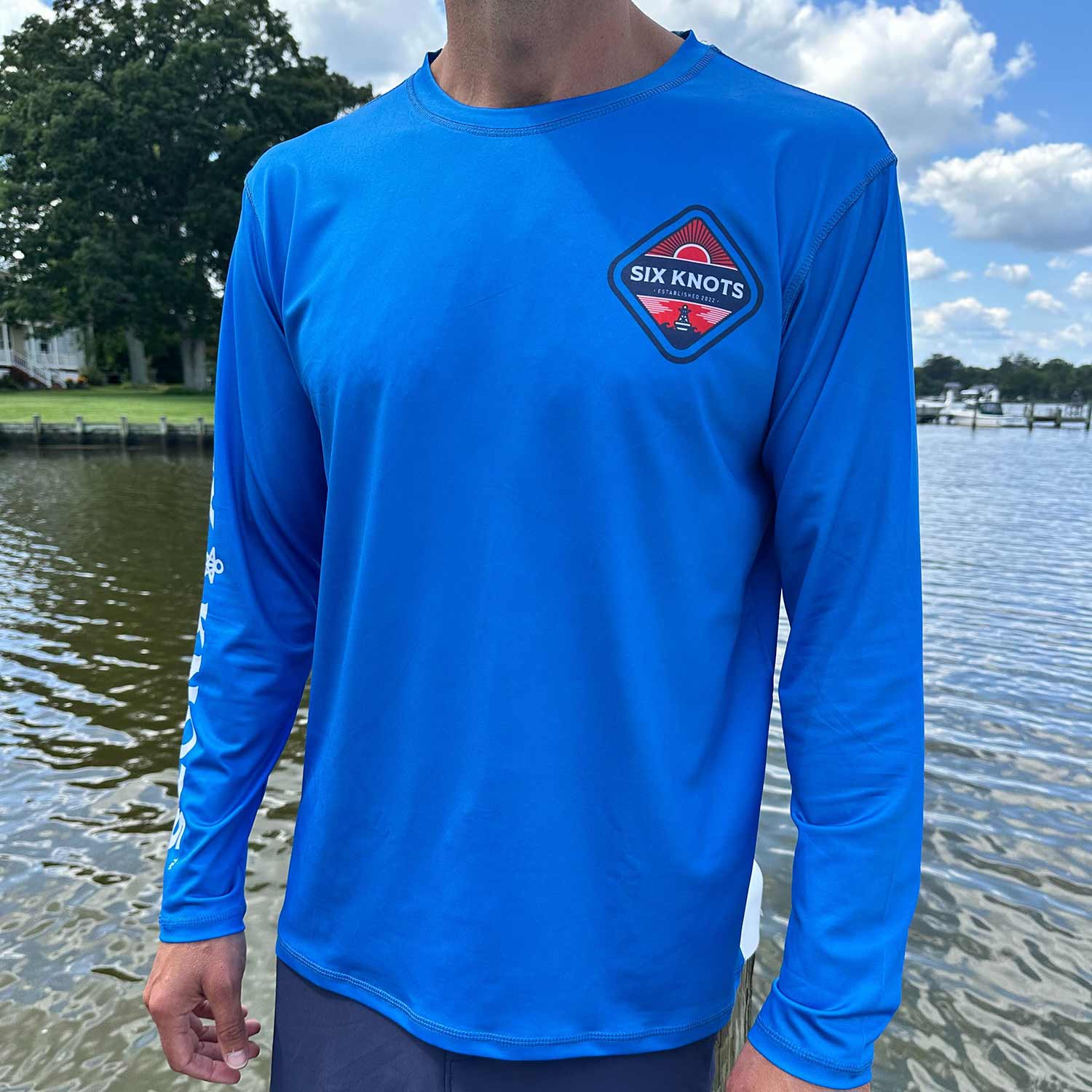 Man standing near the water wearing the Six Knots Red Sky At Night Men’s Performance Crew in vibrant royal blue — lightweight, breathable, and moisture-wicking fishing crewneck shirt designed with UPF sun protection and quick-dry fabric; ideal for saltwater fishing, offshore boating, and rugged all-weather coastal adventures.