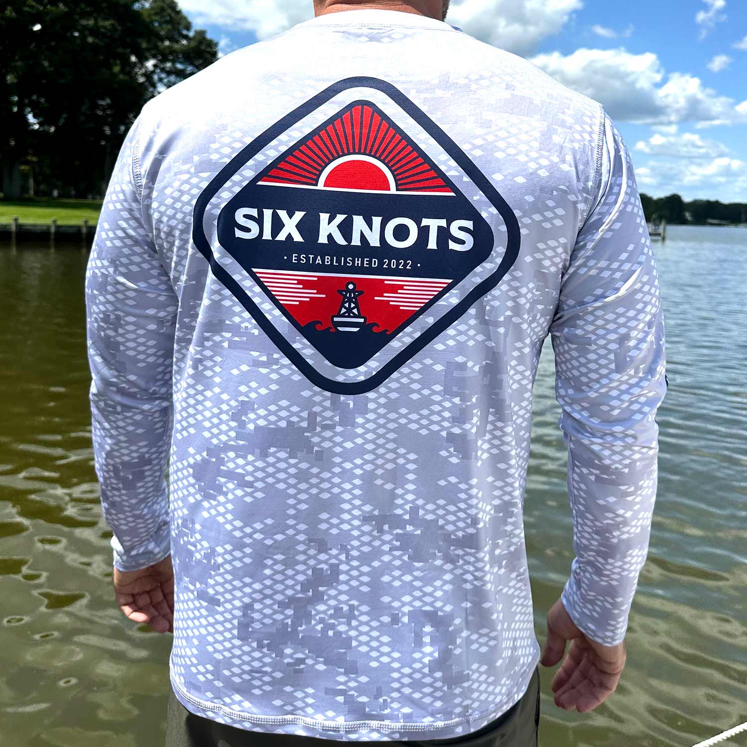 Rear view of man standing near the water wearing the Six Knots Red Sky At Night Men’s Performance Crew in fish scale camo, highlighting the bold red and navy ‘Six Knots Established 2022’ back badge — lightweight, breathable, and moisture-wicking fishing shirt with a digital gray fish scale camo pattern; built with UPF sun protection and quick-dry fabric for saltwater fishing, offshore boating, and all-weather coastal adventures.