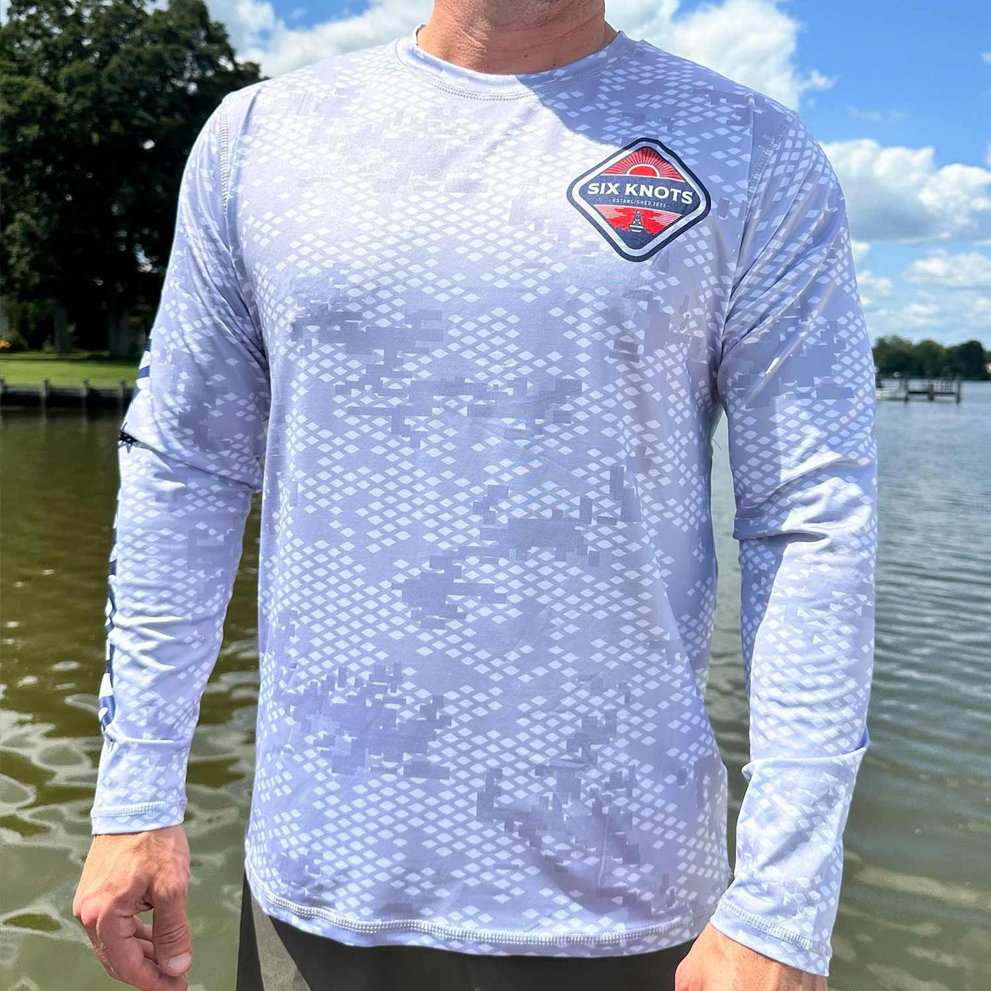 Man standing near the water wearing the Six Knots Red Sky At Night Men’s Performance Crew in fish scale camo — lightweight, breathable, and moisture-wicking crewneck shirt featuring a digital gray camouflage pattern inspired by fish scales; engineered for UPF sun protection and quick-dry comfort, perfect for saltwater fishing, offshore boating, and all-weather coastal adventures.