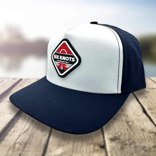 Angled side view of the Six Knots Red Sky At Night Snapback Hat resting on a dock with a blurred marina backdrop, highlighting the breathable mesh back — water-resistant performance fishing hat with a crisp white and navy blue color scheme and a bold front patch that reads ‘Six Knots Performance Apparel Established 2022’ over a red rising sun motif; perfect for saltwater fishing, offshore boating, and all-weather coastal lifestyle wear.