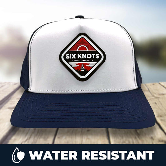 Six Knots Red Sky At Night Snapback Hat resting on a dock with a blurred marina backdrop — water-resistant performance snapback featuring a crisp white and navy blue colorway and a bold front patch reading ‘Six Knots Performance Apparel Established 2022’ over a red rising sun design; ideal for saltwater fishing, offshore boating, and durable all-weather coastal lifestyle wear.