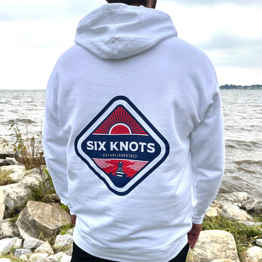 Rear view of man standing by the water wearing the Six Knots Red Sky At Night Classic Hoodie in white, showcasing a bold back graphic with the Six Knots wordmark over a striking red rising sun — ultra-soft, unisex cotton-blend hoodie designed for breathable, all-day comfort. Perfect for cool beach mornings, laid-back seaside outings, and easy layering for oceanfront adventures.