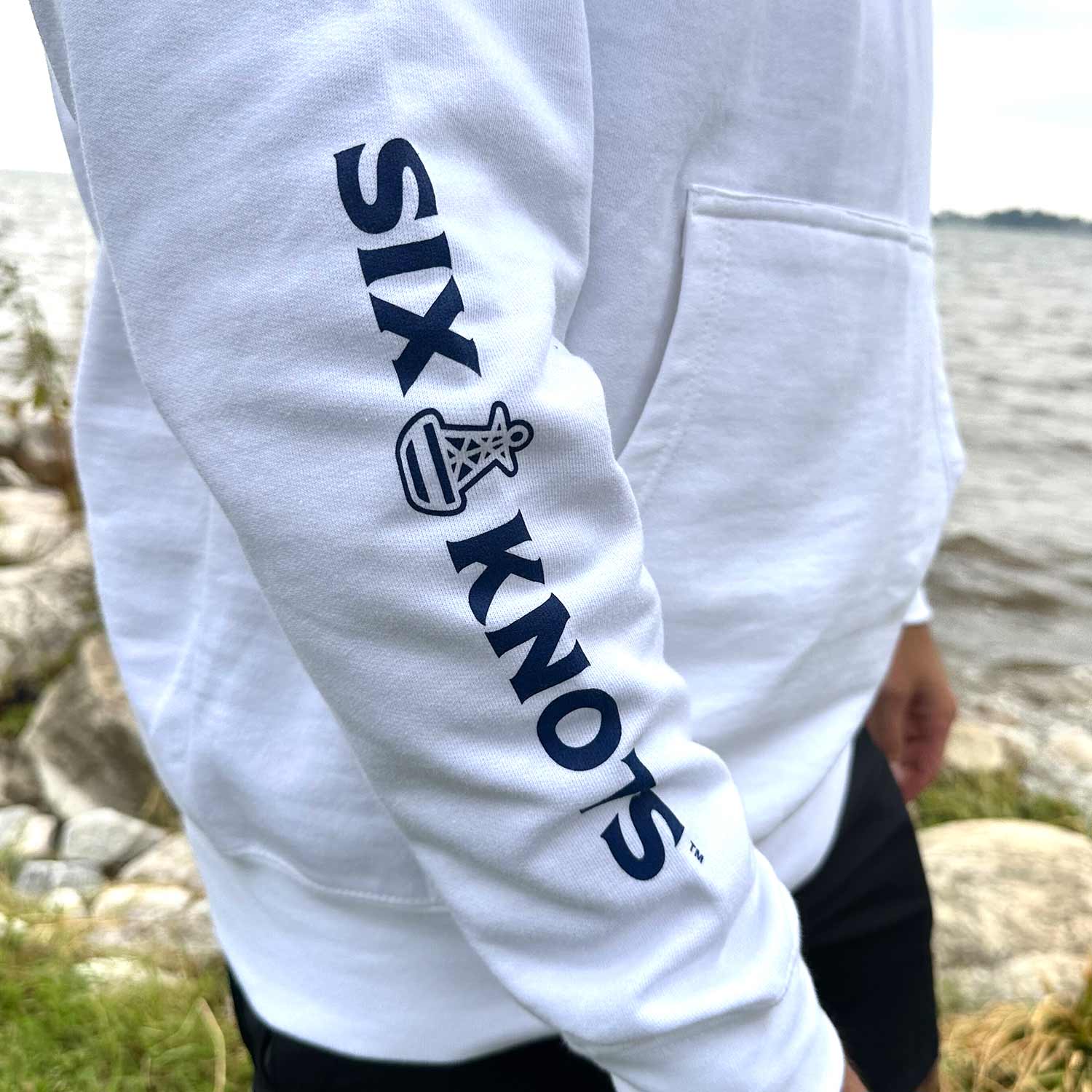 Close-up of the sleeve on the Six Knots Red Sky At Night Classic Hoodie in white, featuring a bold navy Six Knots wordmark running down the arm — ultra-soft, unisex hoodie made from a breathable cotton-blend fabric for all-day coastal comfort. Ideal for cool beach mornings, relaxed seaside outings, and effortless oceanfront layering.