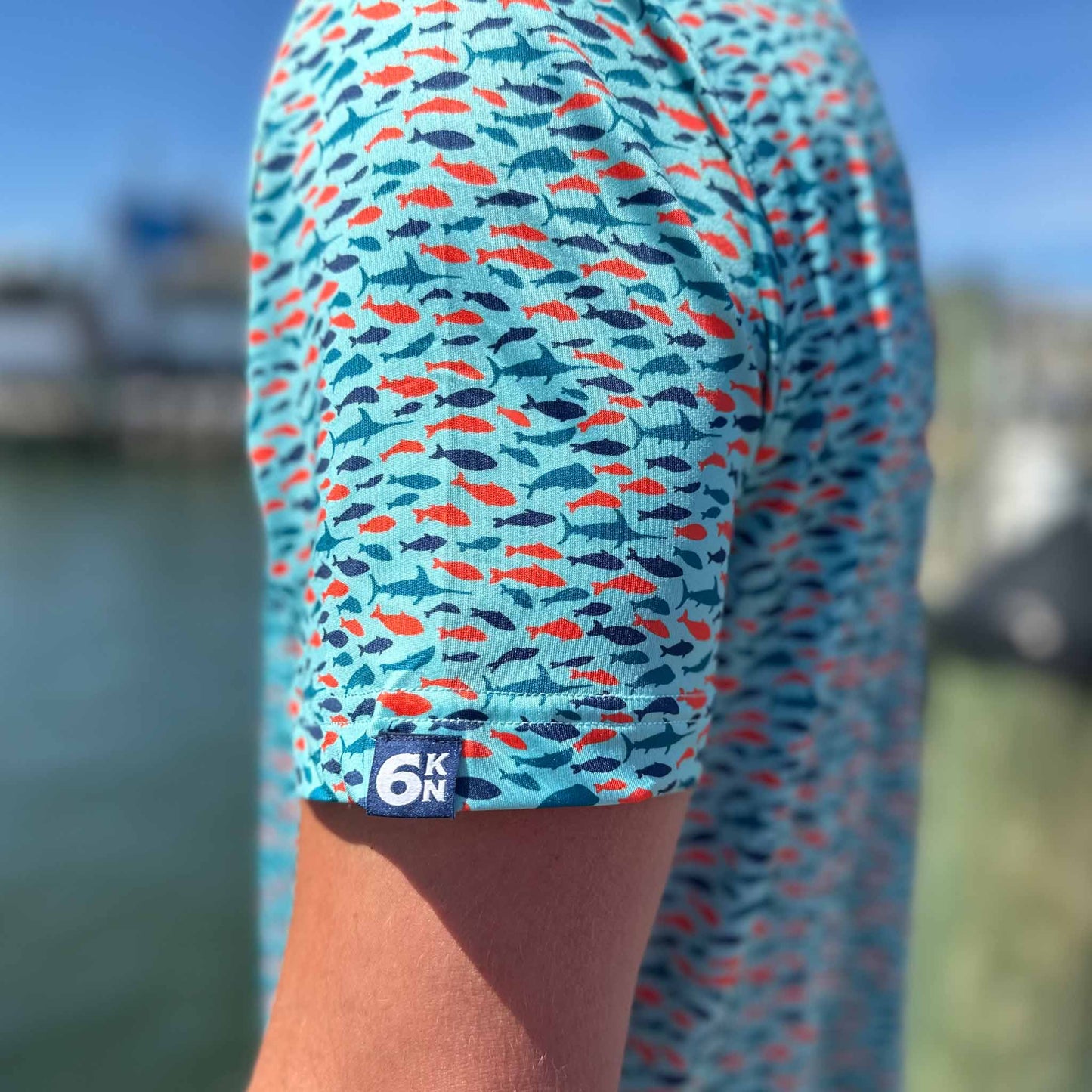 Close-up of Six Knots Schoolie Performance Polo Shirt sleeve featuring the 6KN logo tag and colorful fish pattern — breathable, moisture-wicking men's polo ideal for fishing, boating, and golf