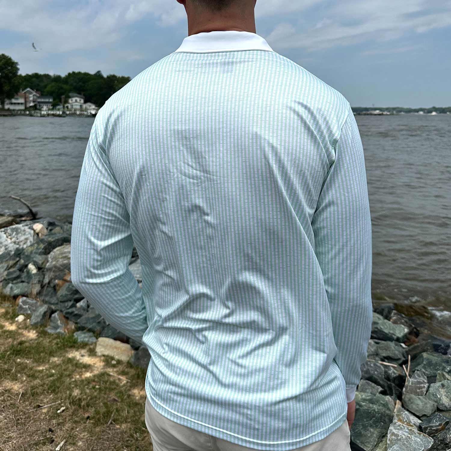Rear view of man standing by coastal rocks and water wearing the Six Knots Seafarer Performance Polo Shirt with mint green seersucker stripes. This lightweight, long-sleeve men's polo features breathable, moisture-wicking fabric and built-in UPF sun protection. Designed for all-day comfort and performance, it's ideal for offshore fishing, boating, seaside travel, and active coastal adventures.