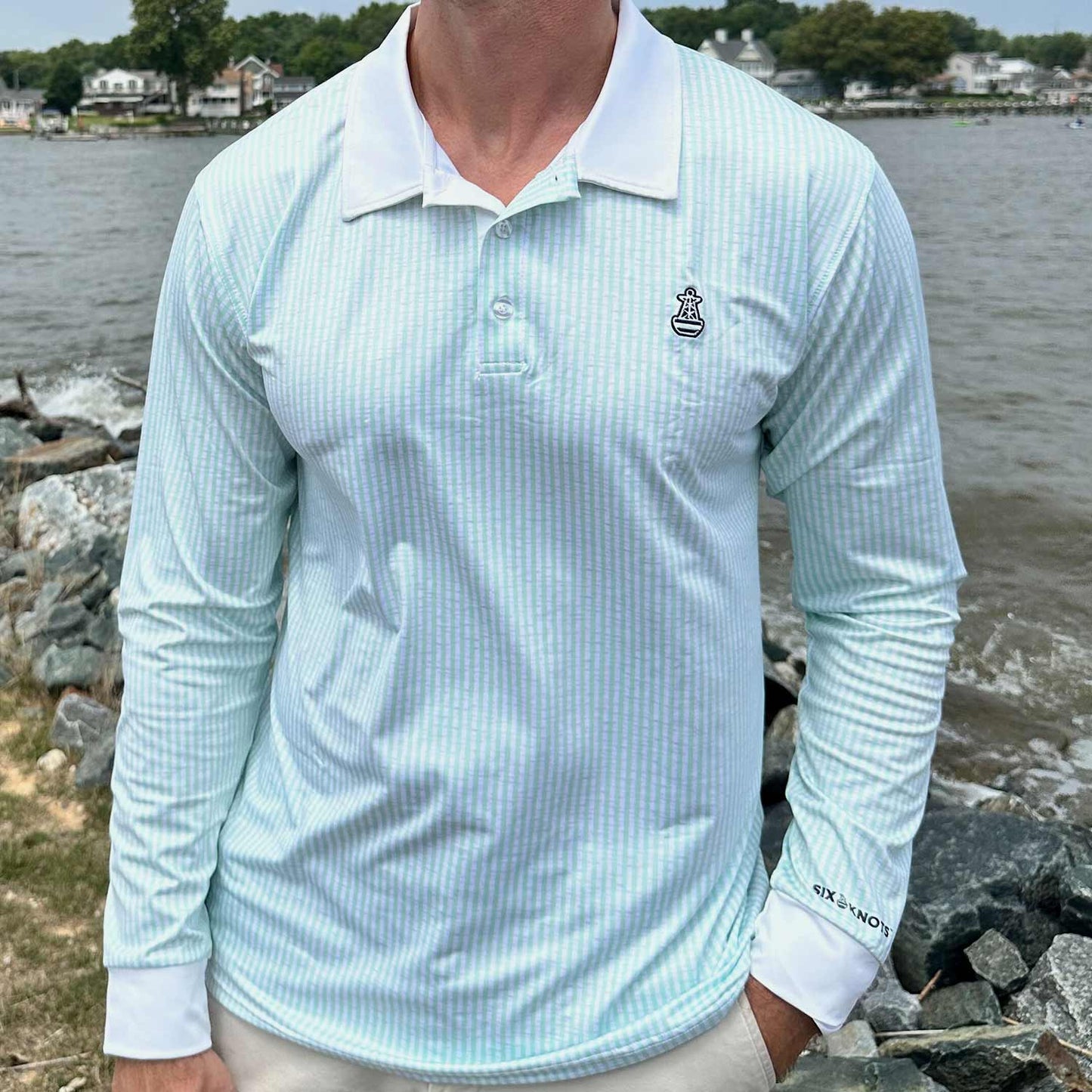 Man standing by coastal rocks and water wearing the Six Knots Seafarer Performance Polo Shirt with mint green seersucker stripes. This lightweight, long-sleeve men's polo is made with moisture-wicking, breathable fabric for superior comfort, UV sun protection, and all-weather performance. Perfect for saltwater fishing, boating, seaside travel, and active coastal lifestyles.