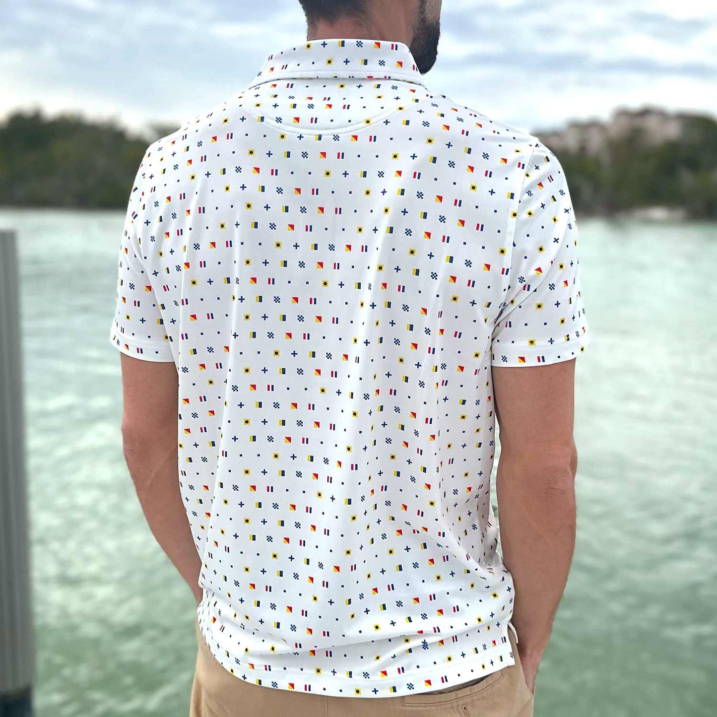 Rear view of a man standing on a boat dock in Florida wearing the Six Knots Signal Flag Performance Polo Shirt. This breathable, lightweight men's polo shirt features a moisture-wicking fabric and a bold nautical signal flag pattern that spells out 'Six Knots.' Ideal for sailing, boating, golfing, beach days, and coastal lifestyle wear