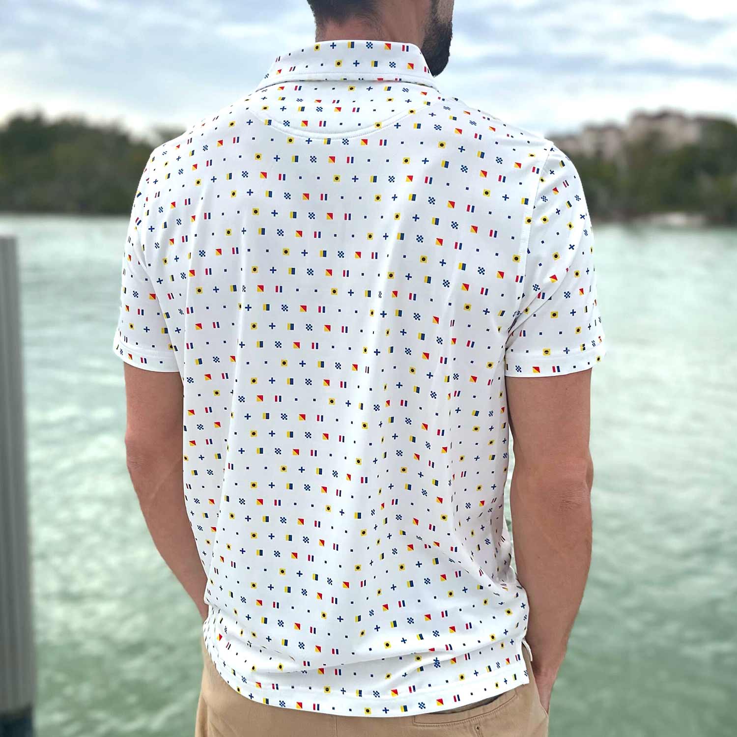 Rear view of a man standing on a boat dock in Florida wearing the Six Knots Signal Flag Performance Polo Shirt. This breathable, lightweight men's polo shirt features a moisture-wicking fabric and a bold nautical signal flag pattern that spells out 'Six Knots.' Ideal for sailing, boating, golfing, beach days, and coastal lifestyle wear