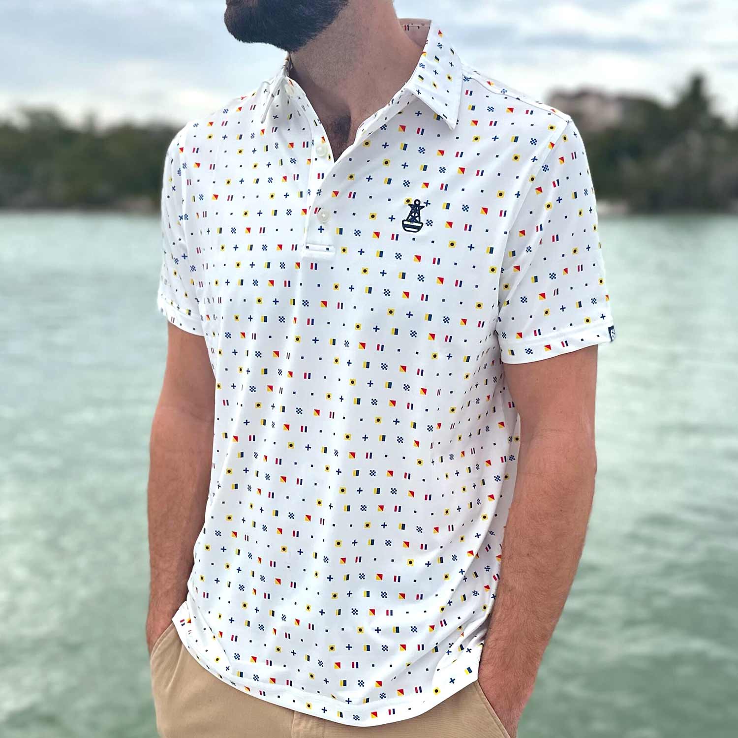 Man standing on a boat dock in Florida wearing the Six Knots Signal Flag Performance Polo Shirt. This lightweight, moisture-wicking men's polo features a repeating nautical signal flag pattern that spells out 'Six Knots' — perfect for sailing, boating, golfing, coastal living, and summer style