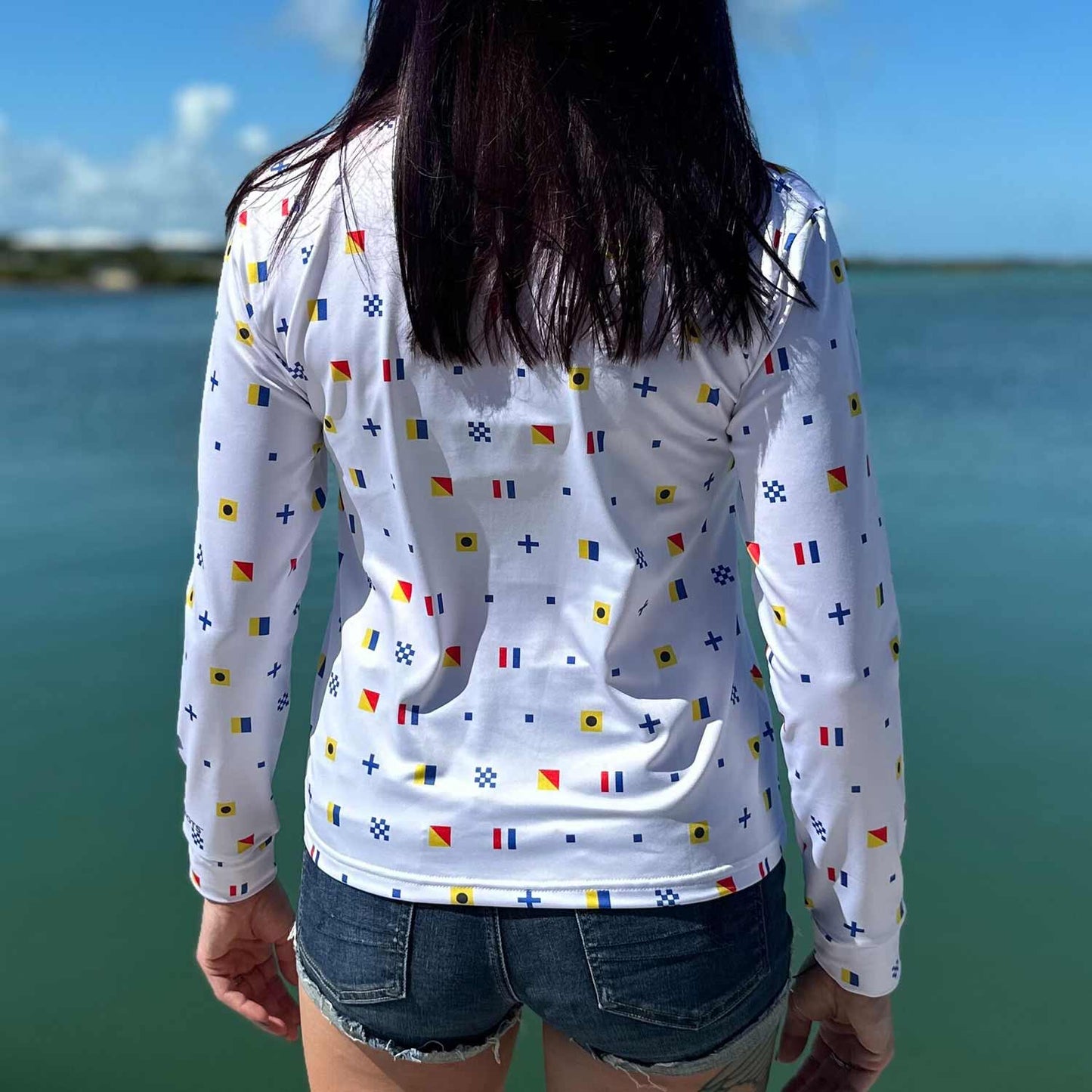 Rear view of woman standing on a marina dock wearing the white Six Knots Signal Flag Women's Performance Quarter Zip, showcasing a repeating nautical signal flag pattern that spells "Six Knots" — lightweight, moisture-wicking, and breathable pullover with UPF sun protection and all-weather comfort, perfect for fishing, boating, sailing, and coastal lifestyle activities
