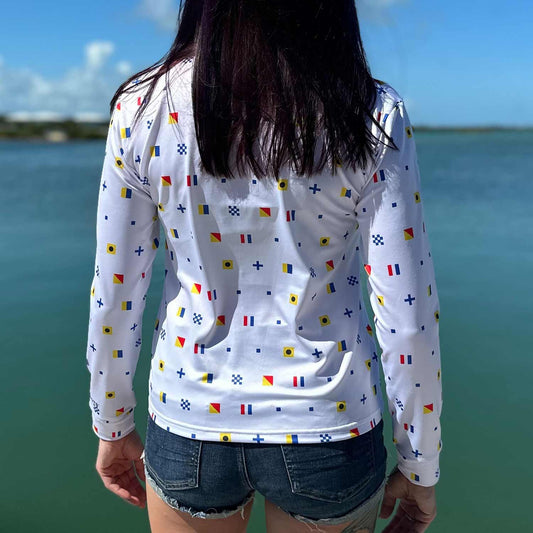 Rear view of woman standing on a marina dock wearing the white Six Knots Signal Flag Women's Performance Quarter Zip, showcasing a repeating nautical signal flag pattern that spells "Six Knots" — lightweight, moisture-wicking, and breathable pullover with UPF sun protection and all-weather comfort, perfect for fishing, boating, sailing, and coastal lifestyle activities