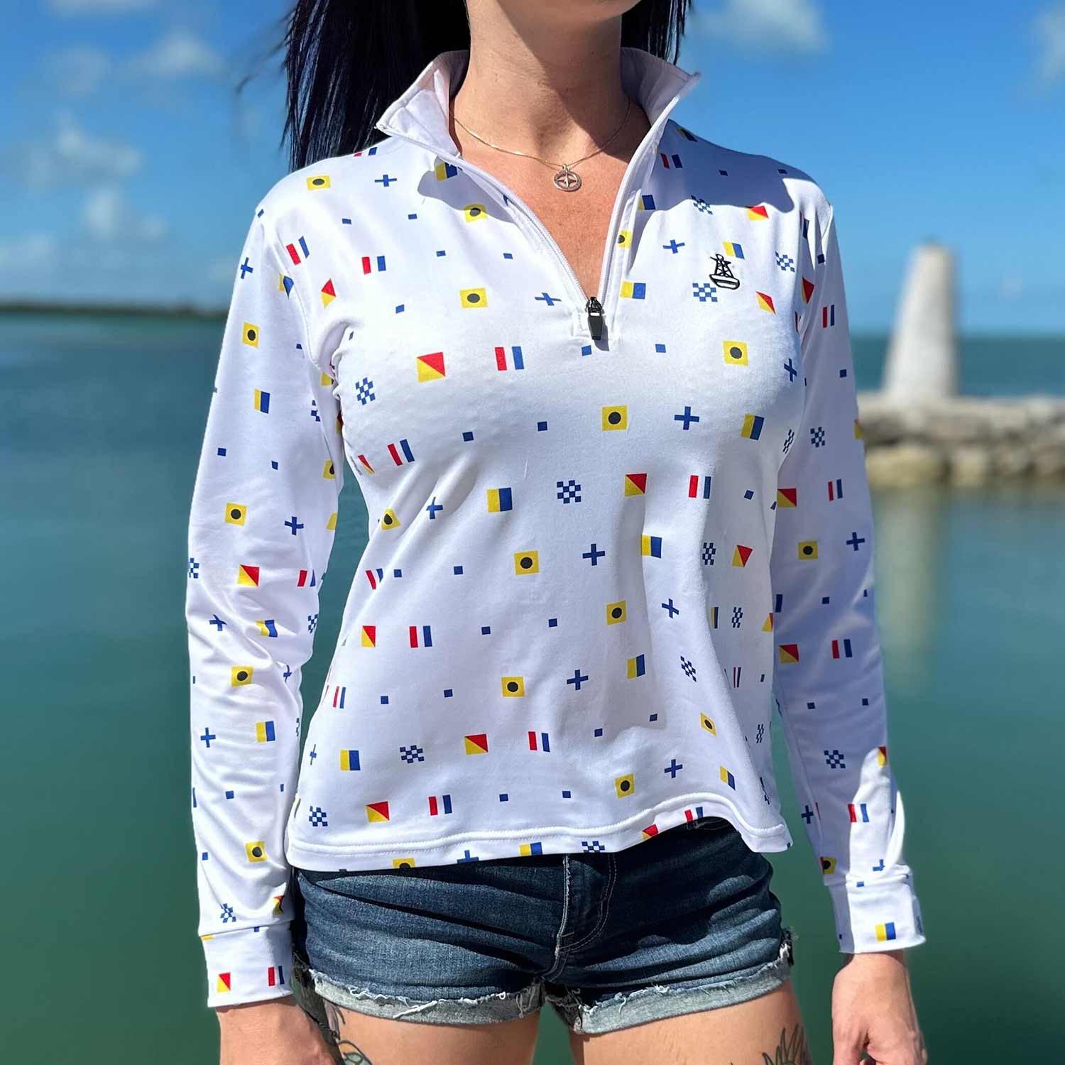 Woman standing on a marina dock wearing the white Six Knots Signal Flag Women's Performance Quarter Zip featuring a repeating nautical signal flag pattern that spells "Six Knots" — lightweight, moisture-wicking pullover with UPF sun protection and all-weather comfort, ideal for fishing, boating, sailing, and coastal adventures