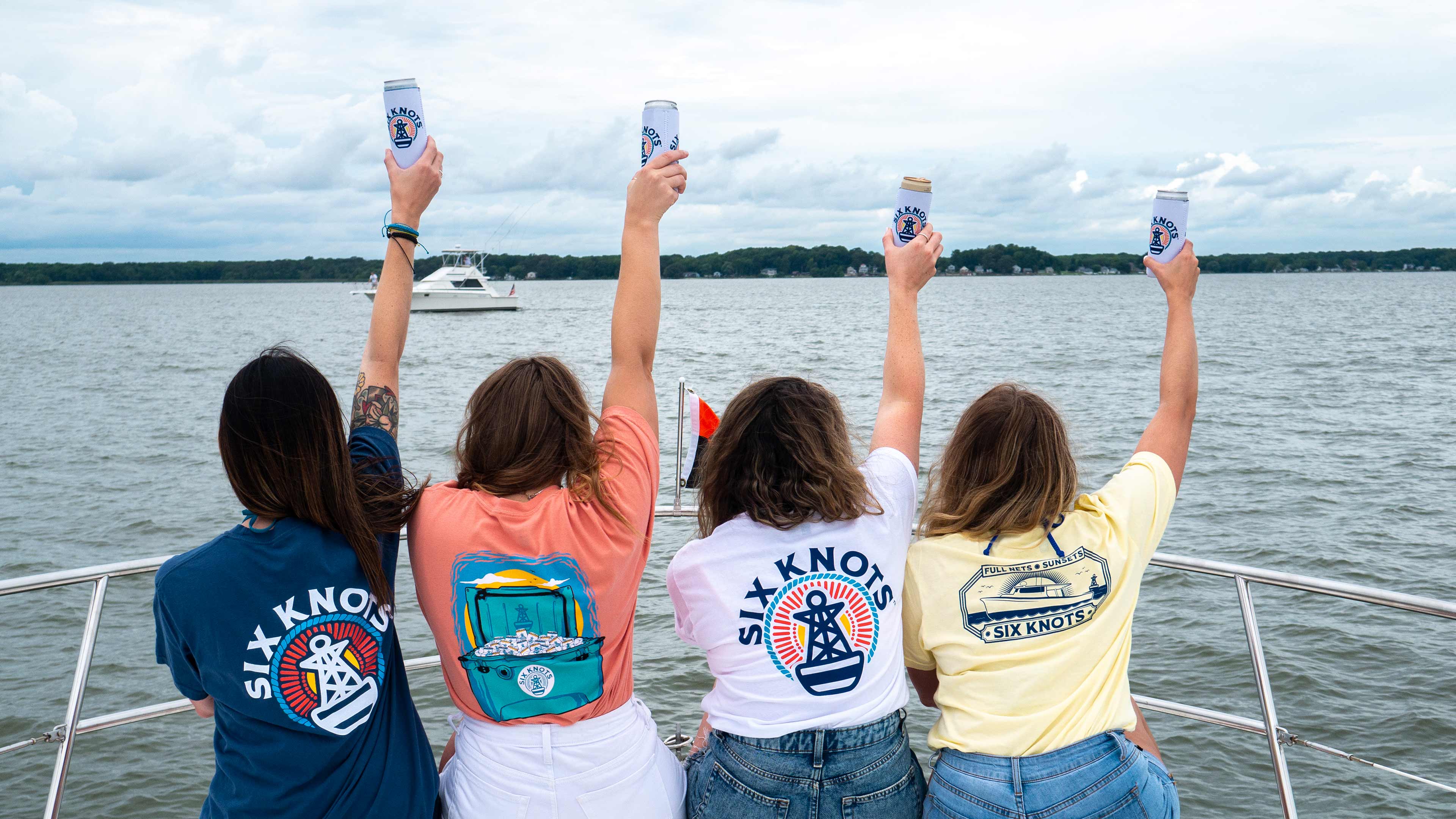 Four girls on a boat wearing shirts from Six Knots and holding seltzers in the sky