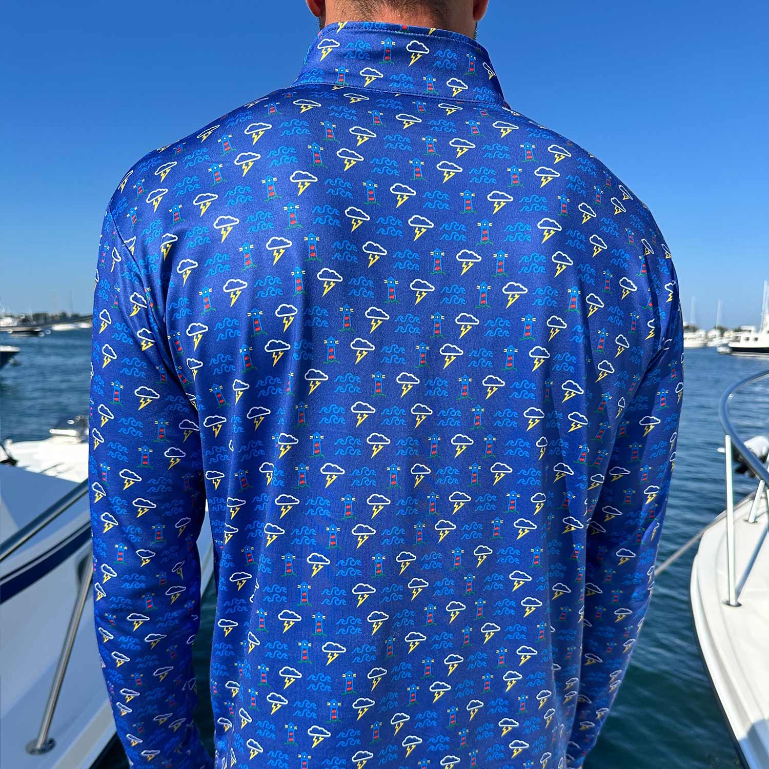 Rear view of man in a marina surrounded by powerboats wearing the Six Knots Storm Front Men’s Performance Quarter Zip — lightweight, breathable, and moisture-wicking blue pullover with an all-over print of lighthouses, ocean waves, and coastal thunderstorms; designed for saltwater fishing, offshore boating, and all-weather coastal adventures.
