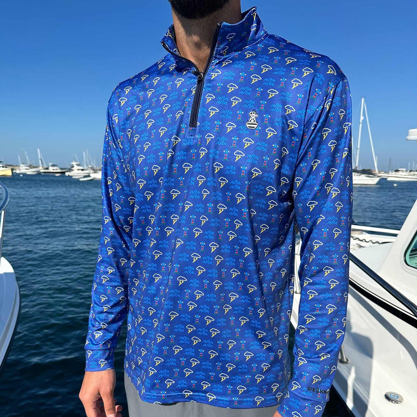 Man standing in a marina surrounded by powerboats wearing the Six Knots Storm Front Men’s Performance Quarter Zip — moisture-wicking, breathable fishing pullover in blue, featuring a repeating pattern of lighthouses, crashing waves, and coastal thunderstorms; ideal for boating, saltwater angling, and all-weather marine adventures.