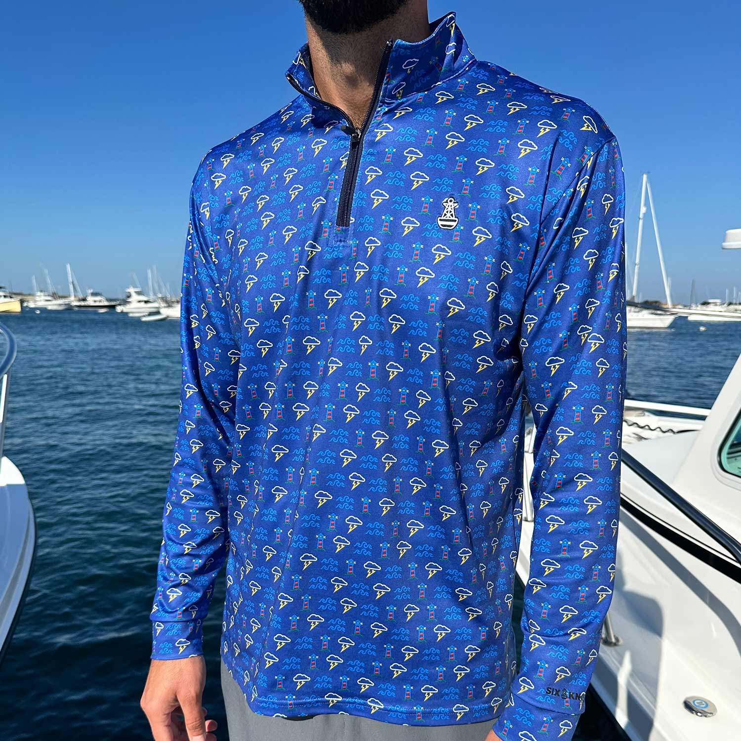 Man standing in a marina surrounded by powerboats wearing the Six Knots Storm Front Men’s Performance Quarter Zip — moisture-wicking, breathable fishing pullover in blue, featuring a repeating pattern of lighthouses, crashing waves, and coastal thunderstorms; ideal for boating, saltwater angling, and all-weather marine adventures.