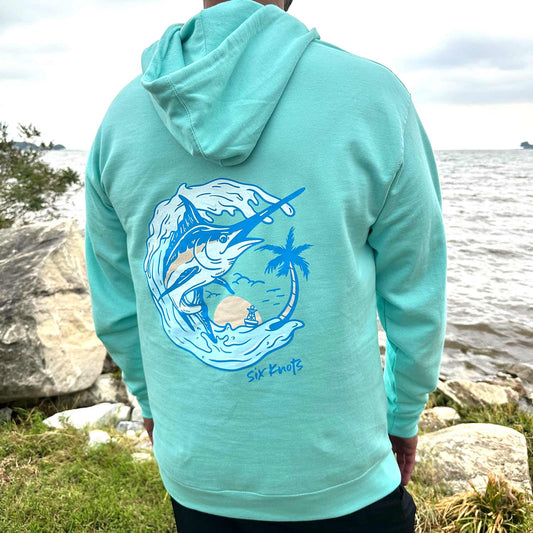 Rear view of man by the water wearing the Six Knots Summer Catch Classic Hoodie in vibrant turquoise, featuring a bold back graphic of a marlin leaping from the water near a tropical island oasis — ultra-soft, unisex cotton-blend hoodie designed for breathable comfort and casual coastal style. Perfect for beach mornings, oceanfront layering, and relaxed seaside adventures.