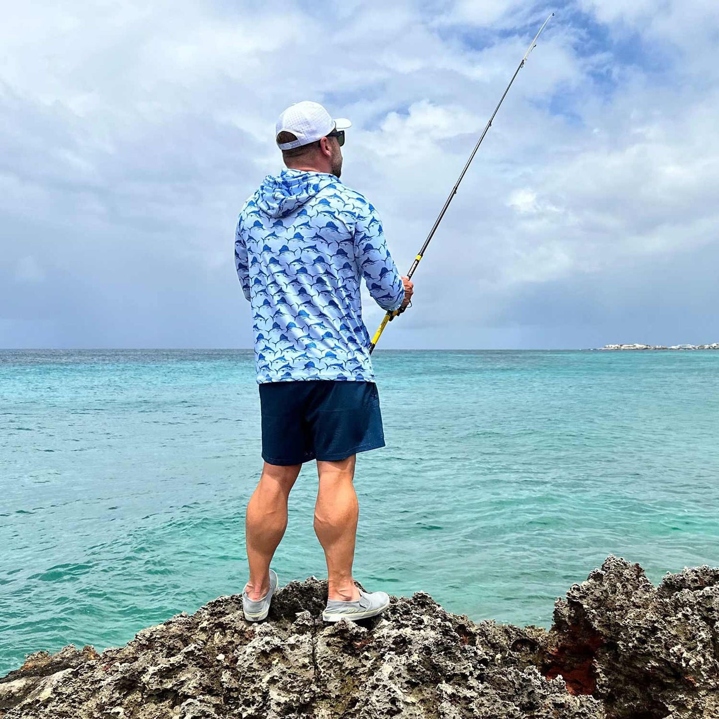 Rear view of man fishing off the coast of St. Maarten wearing a hat and the Six Knots Swords and Sails Performance Hoodie — lightweight, breathable fishing shirt with moisture-wicking fabric and all-over nautical print featuring swordfish and marlins