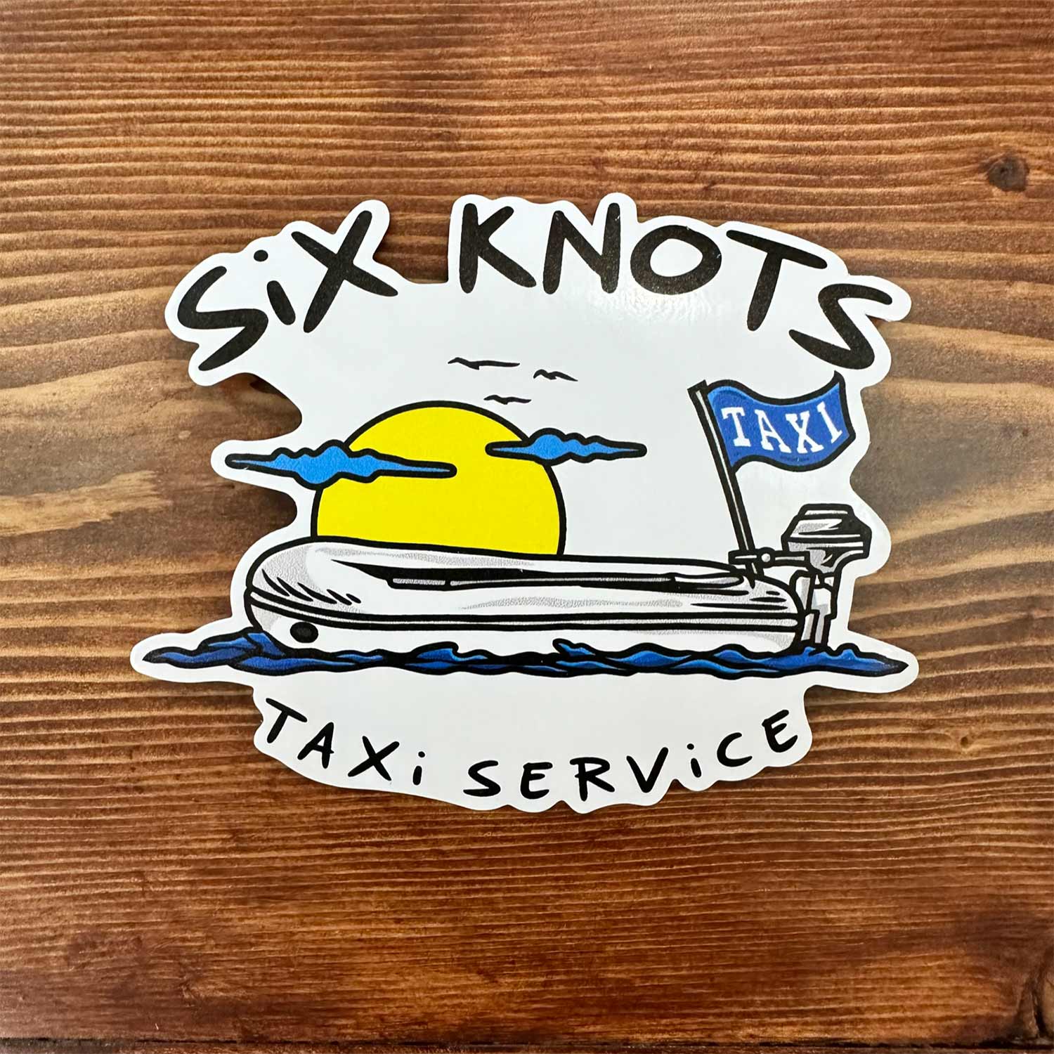 Overhead view of Six Knots "Taxi Service" vinyl sticker featuring an inflatable lifeboat silhouette against a bold yellow sunrise — durable, waterproof decal ideal for coolers, tumblers, tackle boxes, water bottles, and marine gear