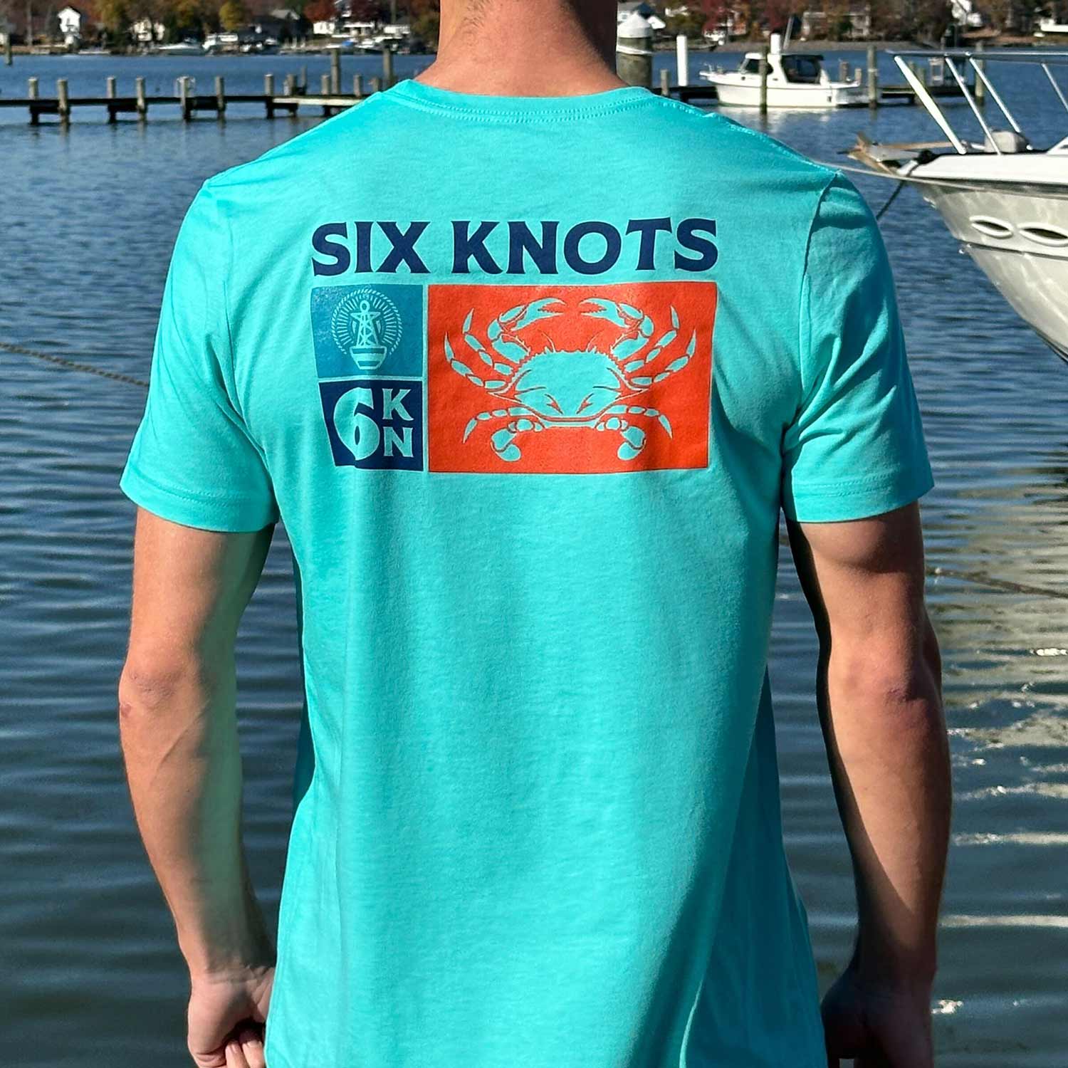 Rear view of man at a Maryland marina wearing the seafoam green Six Knots Maryland Crab Classic T-Shirt. This lightweight, ultra-soft unisex tee is crafted from a breathable cotton-polyester blend and features a bold back graphic of a Maryland blue crab overlaid on a custom flag design made from Six Knots logos. Perfect for everyday wear, beach days, weekend lounging, coastal getaways, casual outings, and laid-back waterfront style.