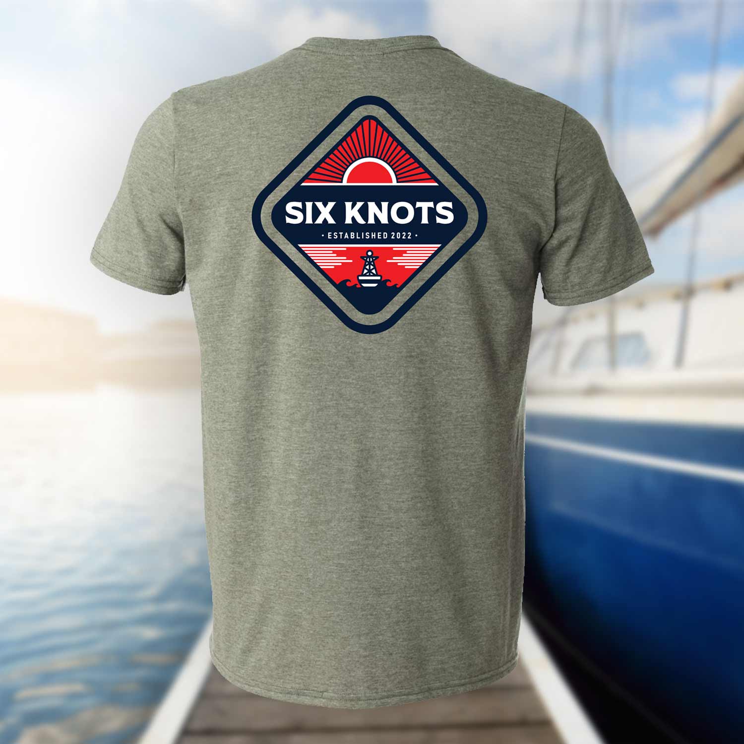 Back view of the Six Knots Red Sky At Night Classic T-Shirt in heather gray set against a blurred marina backdrop, showcasing a bold back graphic with the Six Knots wordmark over a vivid red rising sun. This lightweight, ultra-soft unisex cotton-polyester blend tee offers breathable, all-day comfort—perfect for saltwater fishing, beach mornings, casual coastal outings, and easy oceanfront layering.