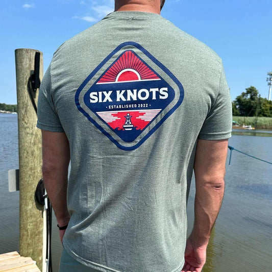 Rear view of man standing by the water wearing the Six Knots Red Sky At Night Classic T-Shirt in heather gray, featuring a bold back graphic with the Six Knots wordmark over a vivid red rising sun. This ultra-soft, unisex cotton-polyester-blend tee is lightweight, breathable, and designed for all-day coastal comfort — ideal for beach mornings, casual seaside outings, and effortless oceanfront layering.