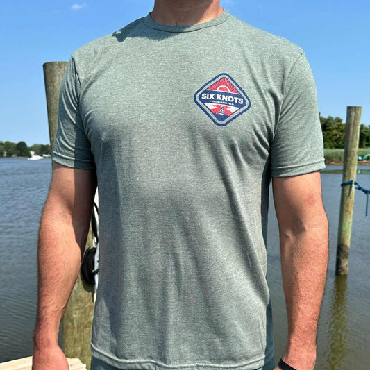Man standing by the water wearing the Six Knots Red Sky At Night Classic T-Shirt in heather gray, showcasing a subtle front chest badge with the Six Knots wordmark over a bold red rising sun graphic. This lightweight, ultra-soft unisex cotton-polyester-blend t-shirt offers breathable, all-day comfort — perfect for saltwater fishing, beach mornings, casual coastal outings, and relaxed oceanfront layering.