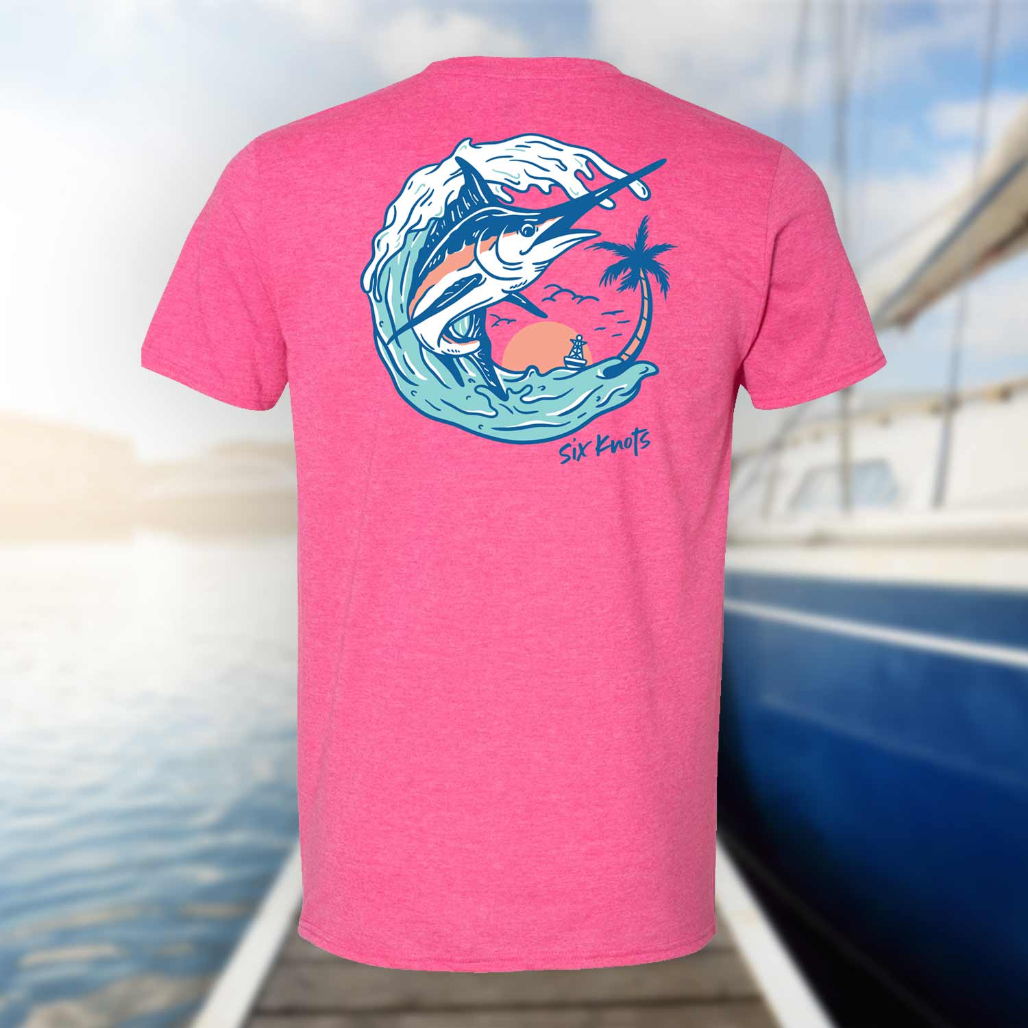 Back view of the Six Knots Summer Catch Classic T-Shirt in vibrant pink, set against a blurred marina backdrop. Features a bold back graphic of a marlin leaping from the water near a tropical oasis. This lightweight, ultra-soft unisex cotton-polyester blend t-shirt is breathable, moisture-wicking, and quick-drying—perfect for saltwater fishing, tropical beach mornings, coastal getaways, and laid-back oceanfront layering.