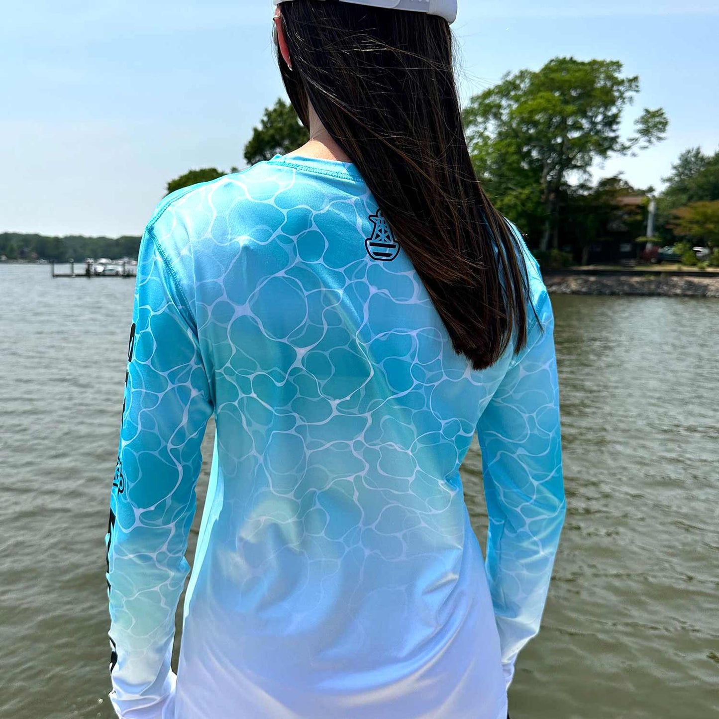 Rear view of woman standing on a powerboat wearing the aqua Six Knots Surf Fisher Women's Performance Crew. This long-sleeve women's fishing shirt features a vibrant water-inspired print, ultra-lightweight moisture-wicking fabric, and UPF 50+ sun protection. Perfect for saltwater fishing, offshore boating, beach adventures, and active coastal living, it delivers breathable comfort and high-performance style on and off the water.