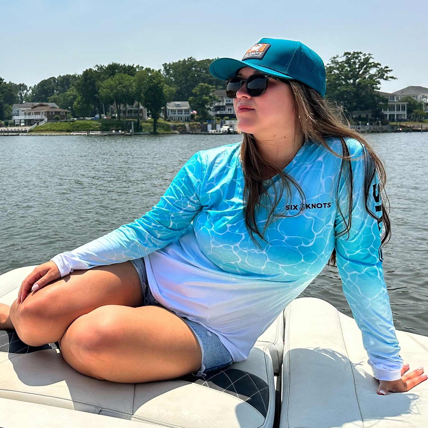 Woman lounging on a powerboat wearing the aqua Six Knots Surf Fisher Women's Performance Crew. This long-sleeve fishing shirt features a unique water-inspired print, lightweight moisture-wicking fabric, and built-in UPF 50+ sun protection. Ideal for saltwater fishing, boating, beach days, and active coastal lifestyles, it delivers breathable comfort and all-day performance on the water.