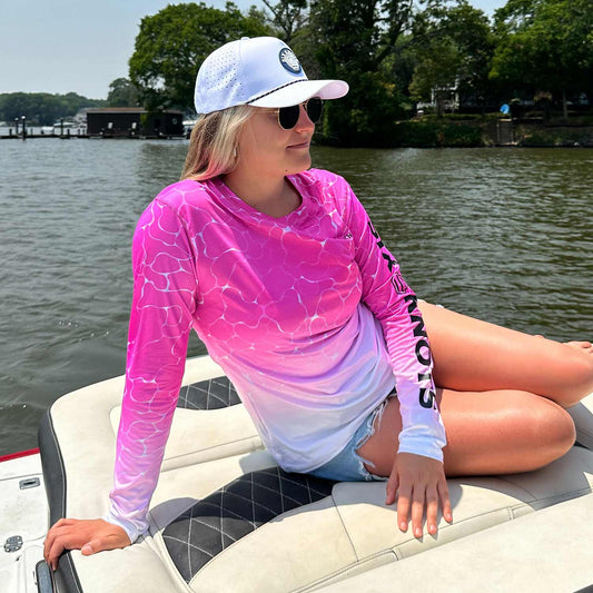 Woman lounging on a powerboat wearing the pink Six Knots Surf Fisher Women's Performance Crew. This long-sleeve fishing shirt features a unique water-inspired print, lightweight moisture-wicking fabric, and built-in UPF 50+ sun protection. Ideal for saltwater fishing, boating, beach days, and active coastal lifestyles, it delivers breathable comfort and all-day performance on the water.