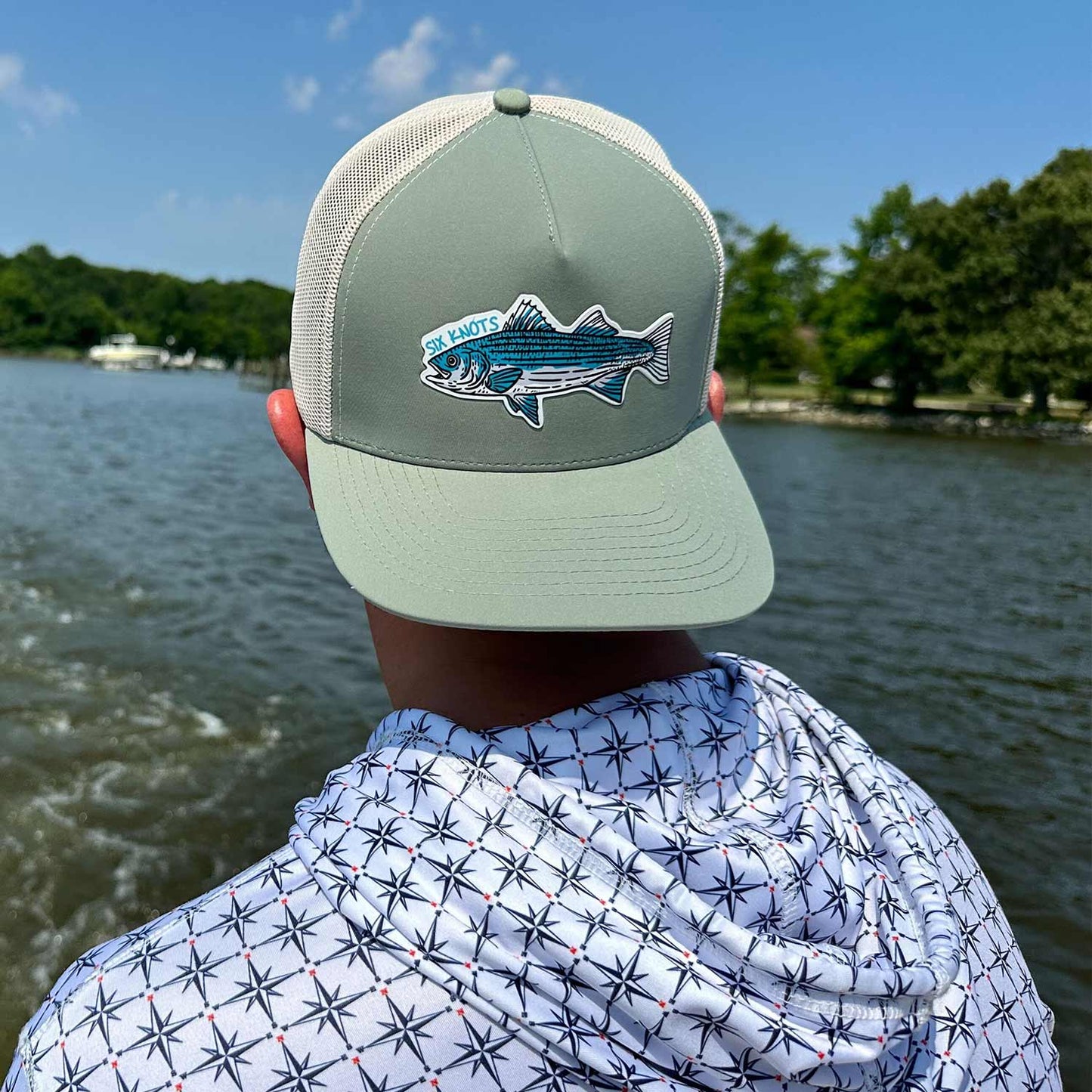 Man sitting on a boat wearing the Six Knots Trophy Season Snapback Hat backwards — water-resistant, breathable performance fishing hat featuring a green and tan colorway and an embroidered striped bass patch. Perfect for saltwater fishing, offshore boating, and rugged everyday wear along the coast.