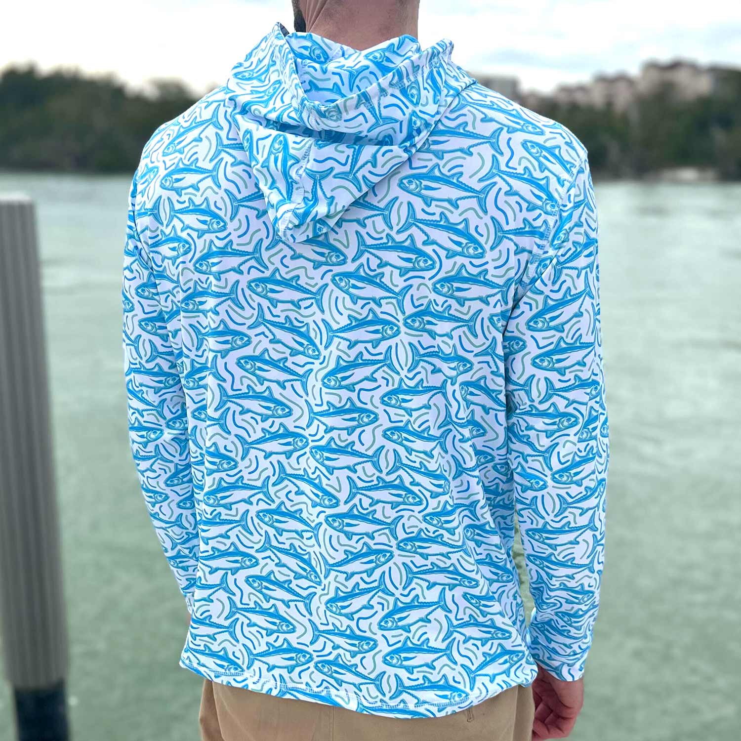 Rear view of man standing on a Florida dock wearing the Six Knots Tuna Time Performance Hoodie — lightweight, moisture-wicking fishing hoodie featuring a bold blue and green tuna pattern with wavy line details; perfect for saltwater fishing, offshore boating, and sunny coastal adventures