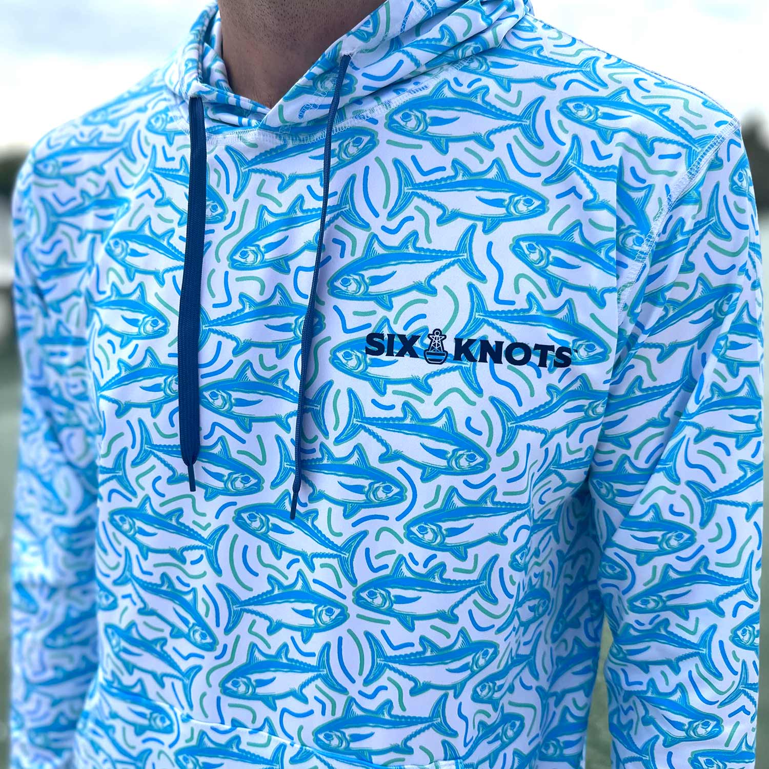 Close-up of the Six Knots Tuna Time Performance Hoodie — breathable, quick-dry fishing hoodie with a vibrant blue and green repeating tuna pattern and wavy line accents; ideal for saltwater fishing, offshore boating, and warm-weather coastal adventures