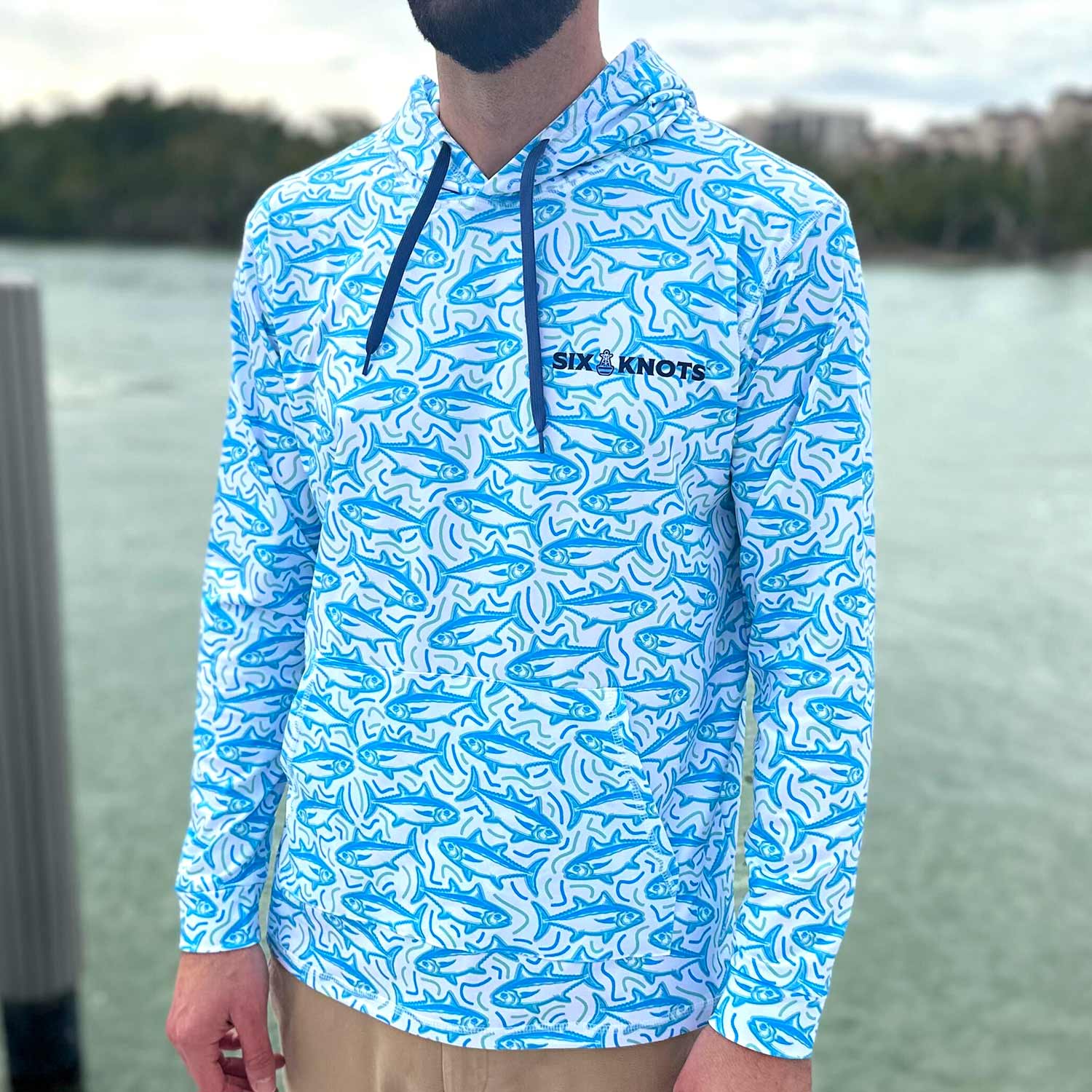 Man standing on a dock in Florida wearing the Six Knots Tuna Time Performance Hoodie — lightweight, moisture-wicking fishing hoodie featuring a vibrant blue and green tuna pattern with wavy line details; ideal for saltwater fishing, boating, and coastal adventures