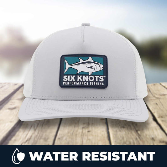 Six Knots Tuna Chaser Snapback Hat resting on a dock with a blurred marina backdrop. This stylish, water-resistant gray snapback features a bold front patch showcasing a tuna and the 'Six Knots Performance Fishing' logo. Designed for durability and coastal performance, it's the perfect hat for saltwater fishing, offshore boating, beach trips, and all-weather outdoor adventures.