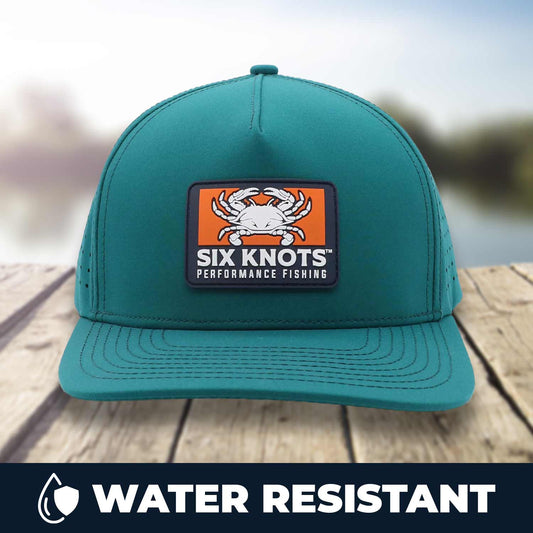 Six Knots Waterman Snapback Hat resting on a dock with a blurred marina backdrop. This eye-catching teal, water-resistant snapback features a bold front patch with a Maryland blue crab and 'Six Knots Performance Fishing' logo. Engineered for coastal durability, breathability, and sun protection, it's the ultimate hat for saltwater fishing, offshore boating, beach outings, and all-weather coastal adventures.