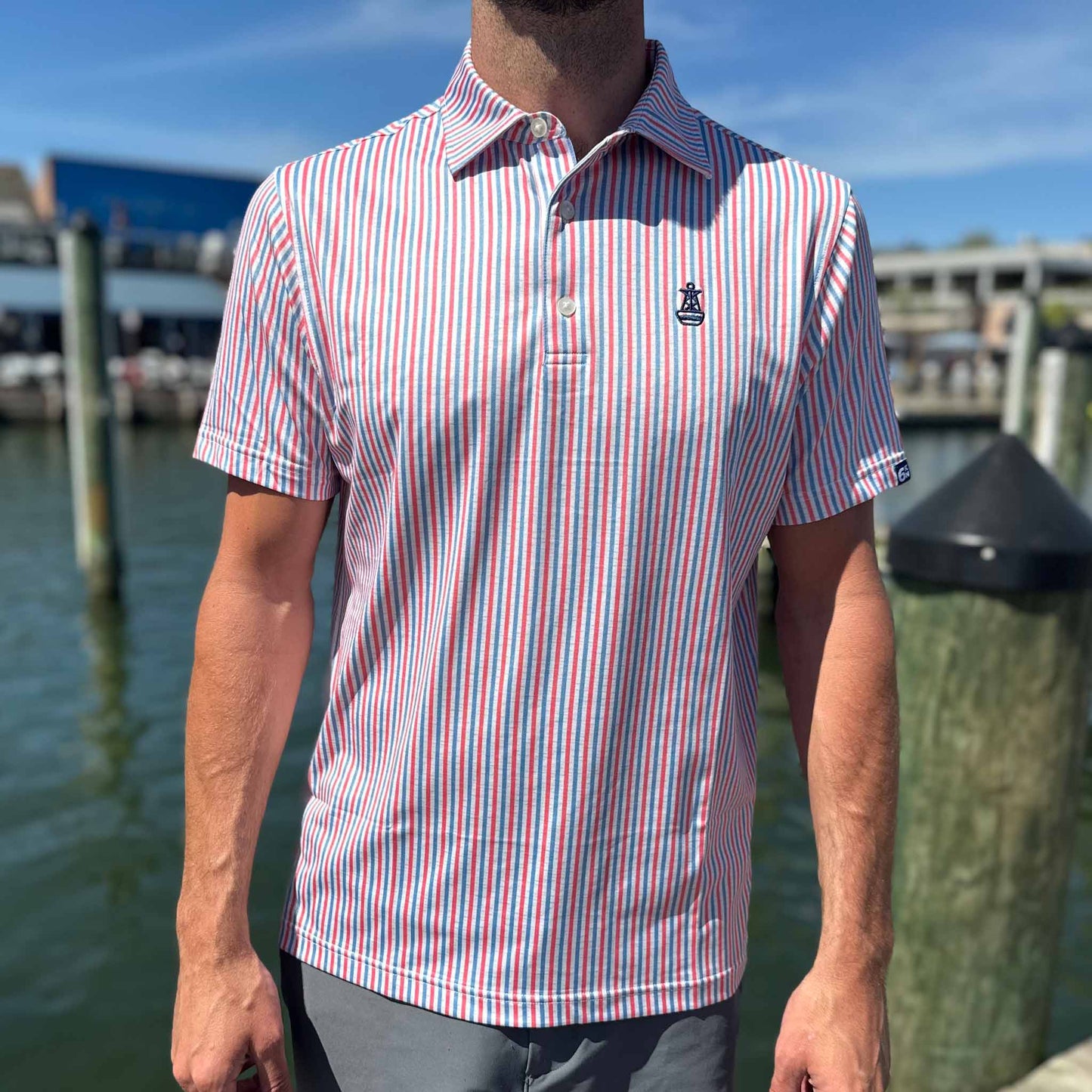 Man wearing the Six Knots All American Performance Polo Shirt on a marina dock, showcasing patriotic red, white, and blue seersucker stripes — breathable, moisture-wicking men's polo perfect for fishing, boating, golf, and summer activities