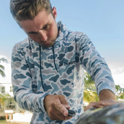 Man filleting a fish at a marina in Fort Lauderdale, Florida while wearing a Six Knots Coastal Camo Performance Hoodie in the Chesapeake pattern — lightweight, moisture-wicking fishing apparel