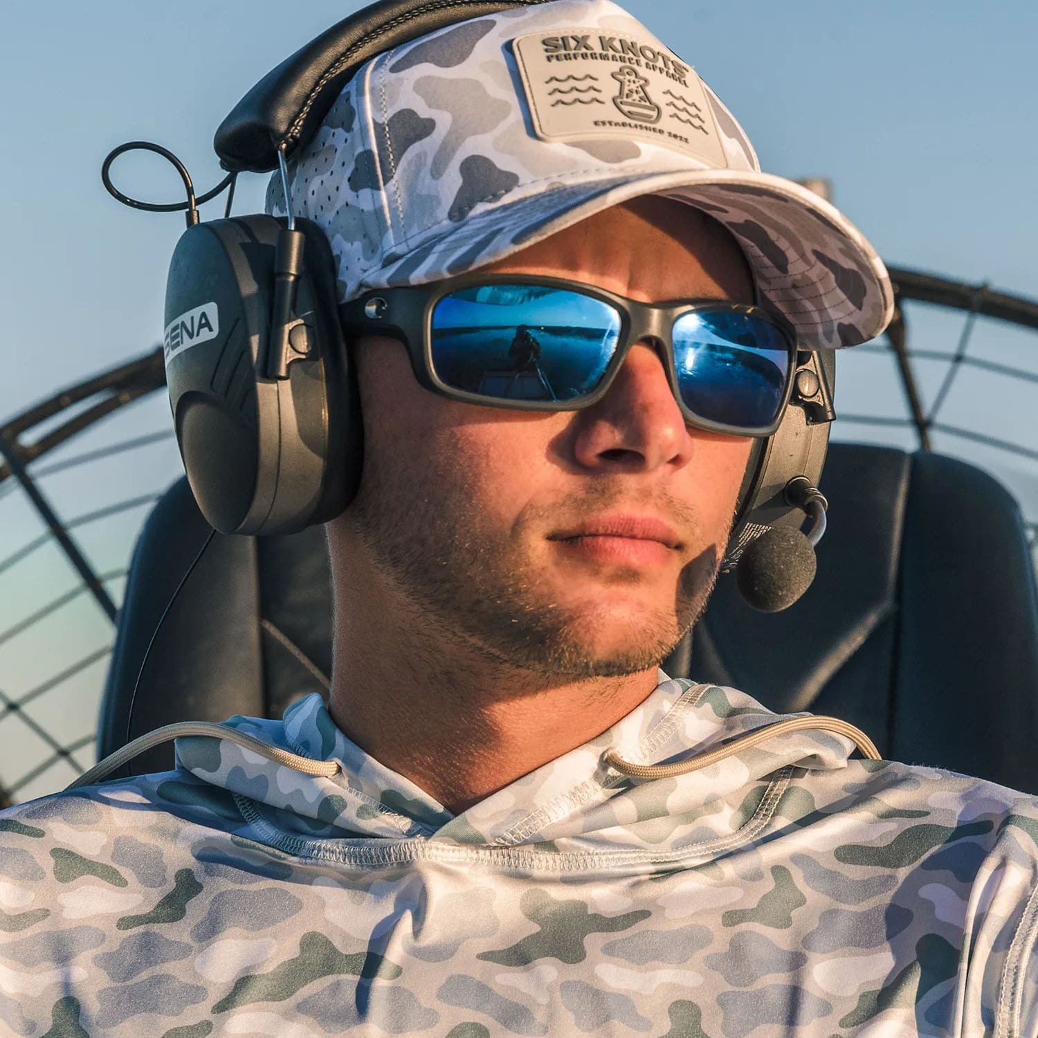 Man airboating in sunglasses, wearing the Six Knots water-resistant Coastal Camo Snapback Hat and Wetlands-pattern Chesapeake Performance Hoodie — lightweight, moisture-wicking boating apparel