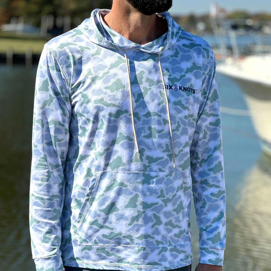 Man standing in a marina wearing the Six Knots Coastal Camo Performance Hoodie in the ‘Wetlands’ colorway — lightweight, breathable fishing hoodie with a blue and green camouflage pattern inspired by coastal wetlands; moisture-wicking and UPF sun-protective apparel ideal for saltwater fishing, boating, and all-weather marine adventures.