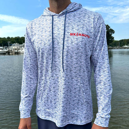 Man standing near the water wearing the Six Knots Feeding Frenzy Performance Hoodie — lightweight, breathable, and moisture-wicking fishing hoodie featuring a striking all-over pattern of feeding sharks and red dots resembling blood in the water. Built for sun protection and comfort, ideal for saltwater fishing, offshore boating, and rugged coastal adventures in any weather.