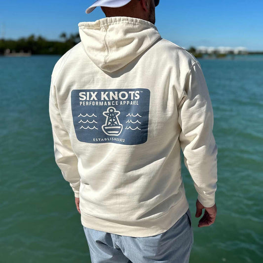 Rear view of man standing by the water wearing the Six Knots Making Waves Classic Hoodie in cream. This ultra-soft, lightweight unisex hoodie features a breathable cotton-blend fabric and relaxed fit for everyday comfort. Ideal for layering on cool coastal mornings, casual weekends, daily wear, travel days, and laid-back seaside style.