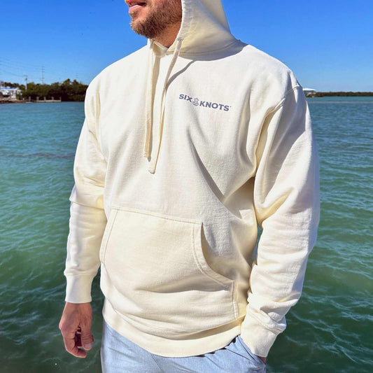 Man standing by the water wearing the Six Knots Making Waves Classic Hoodie in cream. This ultra-soft, lightweight unisex hoodie is made from a breathable premium cotton-blend fabric and designed for all-day comfort and versatility. Perfect for layering on breezy coastal mornings, relaxed weekends, everyday errands, and easygoing travel or leisure wear.