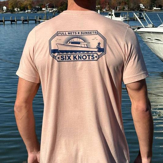 Rear view of man at a Maryland marina wearing the peach Six Knots Perfect Day Classic T-Shirt. This ultra-soft, lightweight unisex tee is crafted from a breathable cotton-polyester blend and features a bold navy blue back graphic of a fishing boat at sunset with the text 'Full Nets, Sunsets, Six Knots.' Ideal for everyday wear, relaxed weekends, beach days, coastal vacations, casual outings, and easygoing waterfront style.