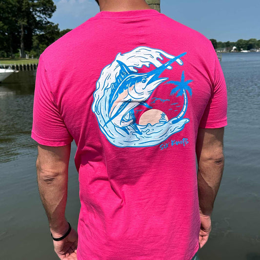 Rear view of man standing by the water wearing the Six Knots Summer Catch Classic T-Shirt in vibrant pink, showcasing a bold back graphic of a marlin jumping from the water in front of a tropical oasis. This lightweight, ultra-soft unisex cotton-polyester blend tee is breathable and moisture-wicking — perfect for everyday wear, weekend lounging, travel days, casual outings, tropical beach mornings, and relaxed seaside layering.