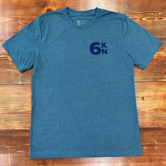 Front view of the deep teal Six Knots Weekender Classic T-Shirt displayed on a wooden table. This ultra-soft, lightweight unisex tee is made from a breathable cotton-polyester blend and features a subtle 6KN wordmark on the left chest. Ideal for everyday wear, weekend lounging, beach vacations, coastal getaways, casual outings, and relaxed waterfront living.