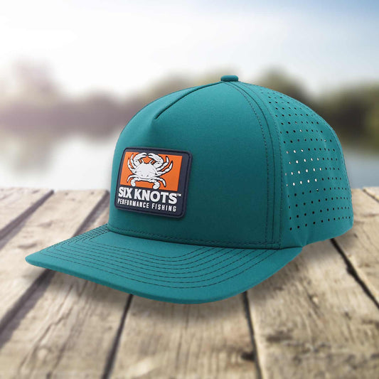 Angled side view of Six Knots Waterman Snapback Hat resting on a dock with a blurred marina backdrop. This eye-catching teal, water-resistant snapback features a bold front patch with a Maryland blue crab and 'Six Knots Performance Fishing' logo. Engineered for coastal durability, breathability, and sun protection, it's the ultimate hat for saltwater fishing, offshore boating, beach outings, and all-weather coastal adventures.